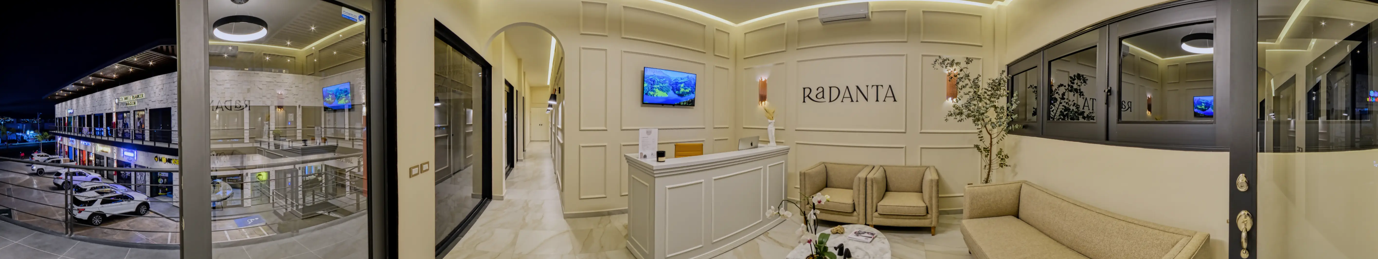 Panorama of the Radanta reception: the glass doors to the walkway, the arch of the hallway to the treatment rooms, the white moulded front desk under the sign and linen armchairs beside an olive tree.